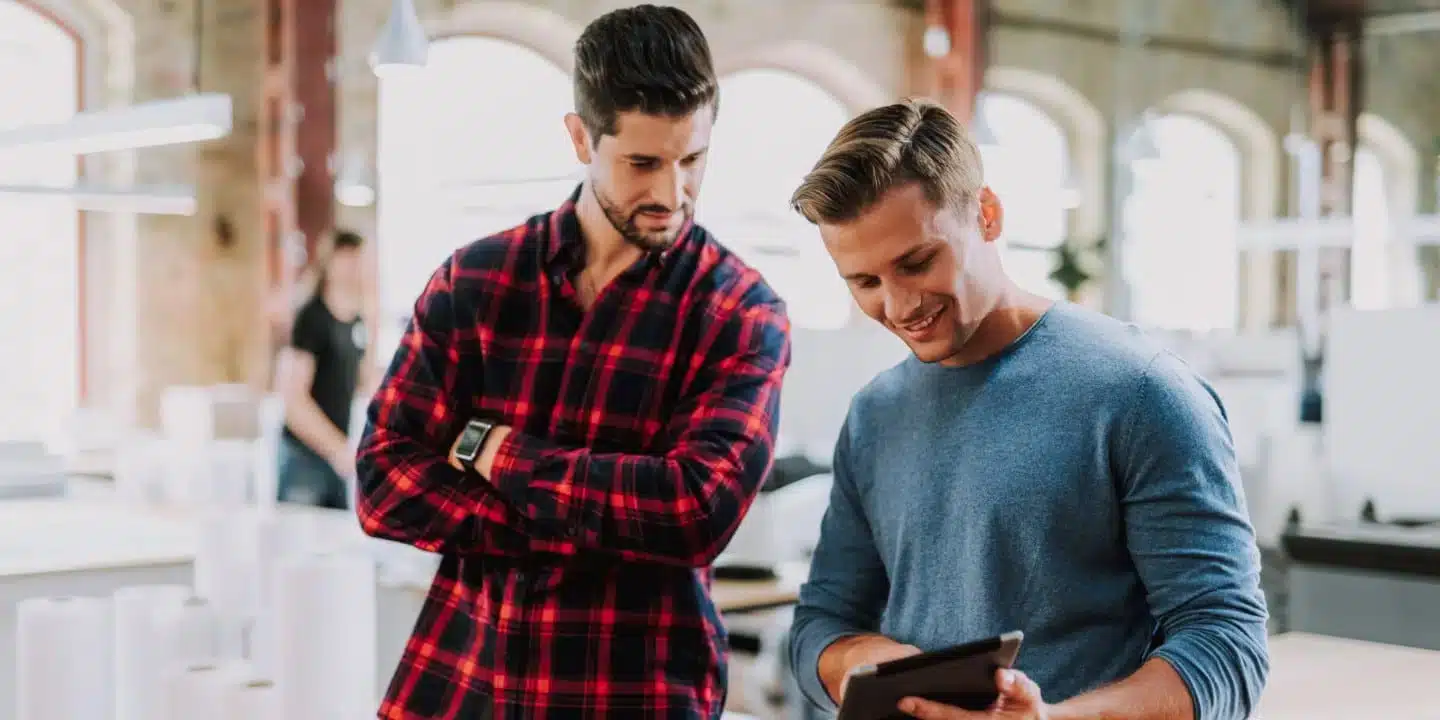 Zwei Männer stehen in einem modernen Büro, einer hält ein Tablet in der Hand, der andere verschränkt die Arme und diskutiert mit seinem PLM-Partner über Lösungen.