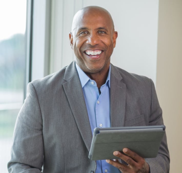 A man in a gray suit holding a tablet stands near a window, smiling at the camera, ready to discuss the PLM idea to cash process.