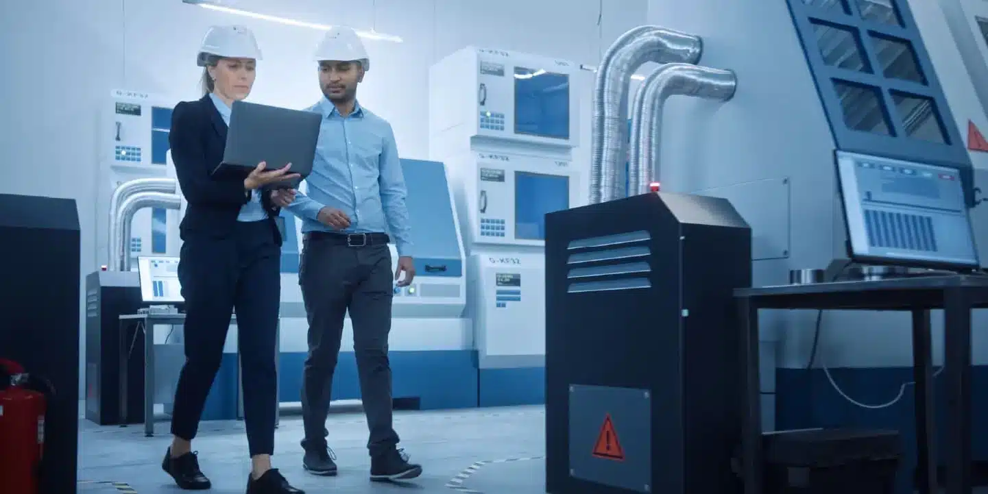 Zwei Ingenieure, die Schutzhelme tragen, überprüfen Details der Produktdokumentenverwaltung auf einem Laptop, während sie durch eine moderne Industrieanlage mit Maschinen und Schalttafeln gehen.