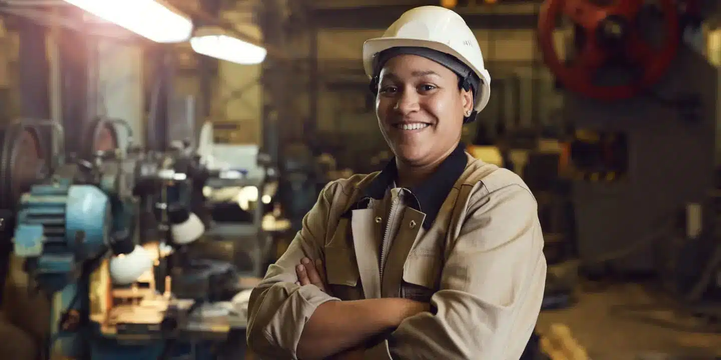 A person wearing a white hard hat and work uniform stands in a factory, smiling with arms crossed. Industrial equipment and BOM Management documents are visible in the background.