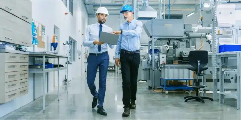 Zwei Männer mit Schutzhelmen gehen durch eine Industrieanlage und diskutieren an einem Laptop über PLM-Produktentwicklung; im Hintergrund sind Maschinen und Anlagen zu sehen.