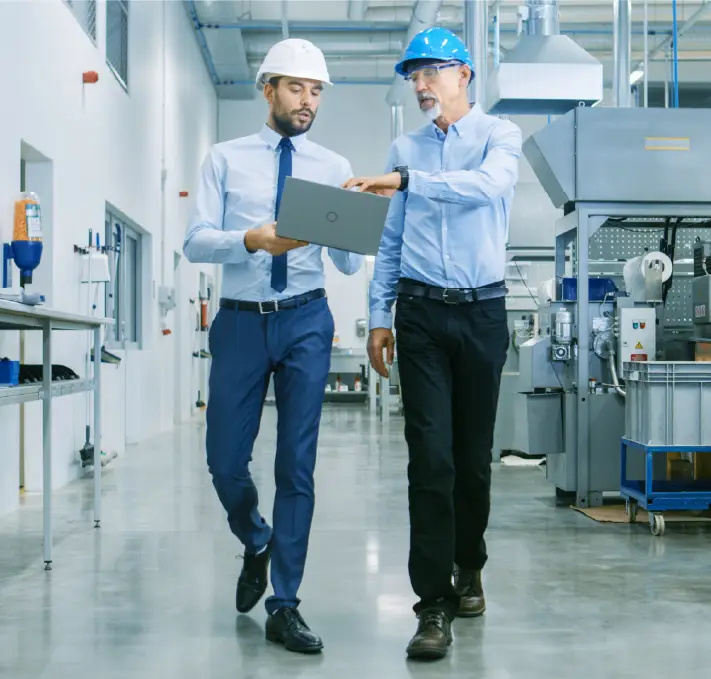 Two men in safety helmets and business attire walk through a factory, examining a laptop and discussing PLM product development in an industrial setting.