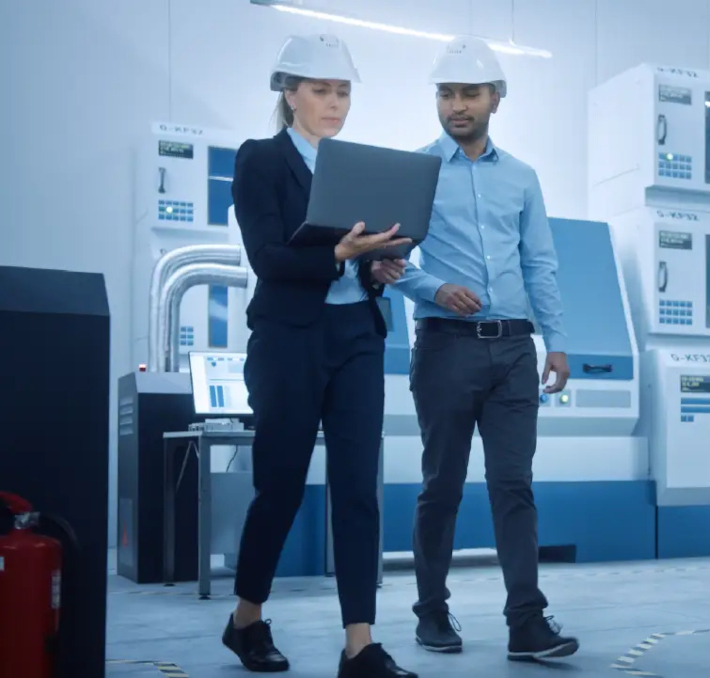 Two engineers wearing hard hats walk through a modern industrial facility; one is holding a laptop, reviewing product document management details.