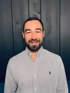 A man with short dark hair and a beard, wearing a light grey button-up shirt, stands in front of a dark paneled wall, smiling at the camera—reflecting confidence in gaining a competitive advantage with PLM.