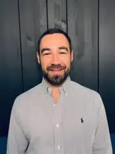 A man with short dark hair and a beard, wearing a light grey button-up shirt, stands in front of a dark paneled wall, smiling at the camera—reflecting confidence in gaining a competitive advantage with PLM.