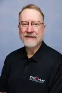 A middle-aged man with glasses and a beard, wearing a black collared shirt with the Creative Security and Design logo, poses in front of a plain light blue background, showcasing their competitive advantage with PLM expertise.