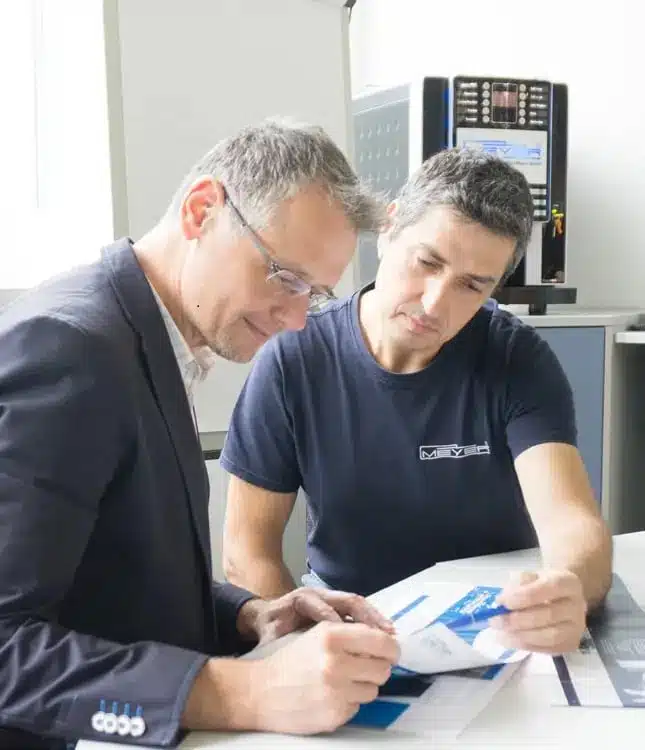Herbert Meyer and a colleague sit at a desk reviewing printed documents together in a brightly lit office setting.