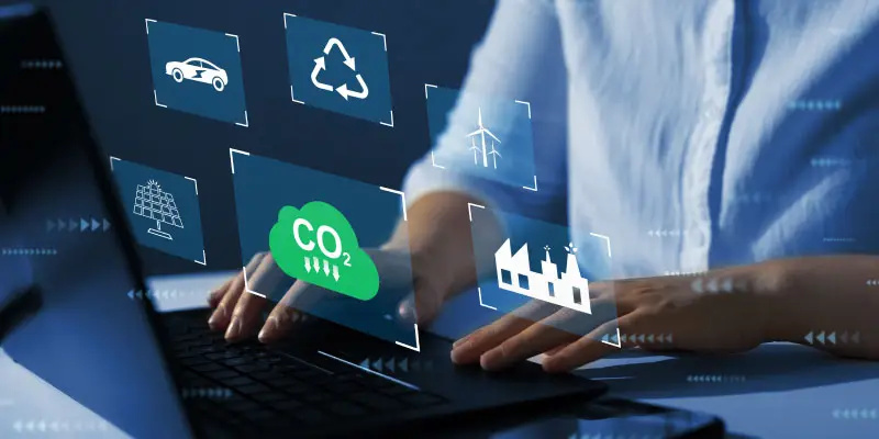 A person types on a laptop, with digital icons for CO2, recycling, solar, wind, electric cars, and factories appearing as virtual graphics above the keyboard.