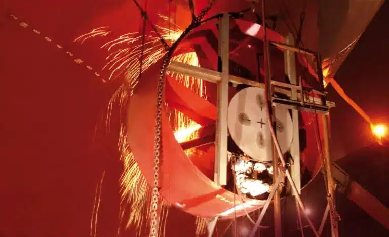Workers welding inside a large circular metal structure at Becker Marine Systems, with bright sparks flying and chains hanging around the work area.
