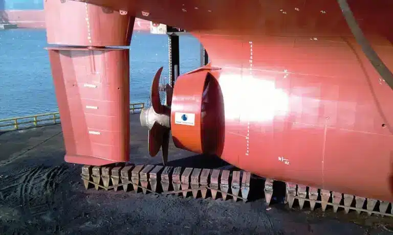 Close-up view of a ship’s propeller and rudder assembly, featuring Becker Marine Systems technology, as the vessel is docked on land near water.