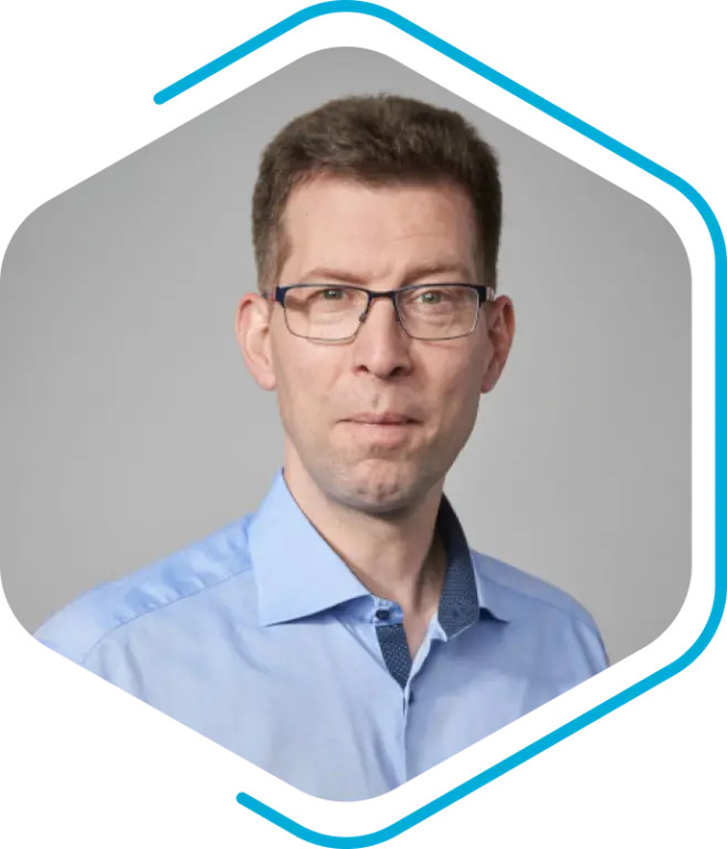 A man with short brown hair and glasses, wearing a light blue collared shirt, poses in front of a plain gray background within a hexagonal frame, representing Becker Marine Systems.