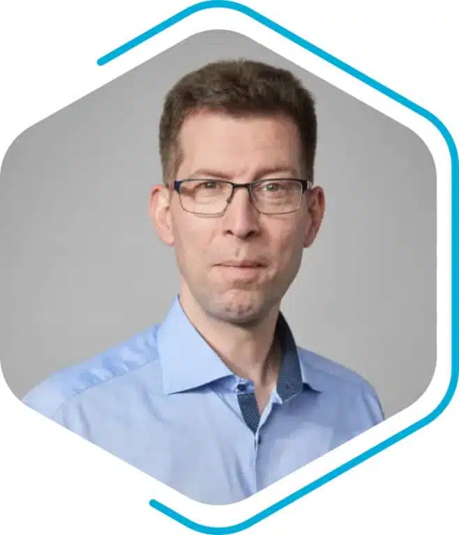 A man with short brown hair and glasses, wearing a light blue collared shirt, poses in front of a plain gray background within a hexagonal frame, representing Becker Marine Systems.