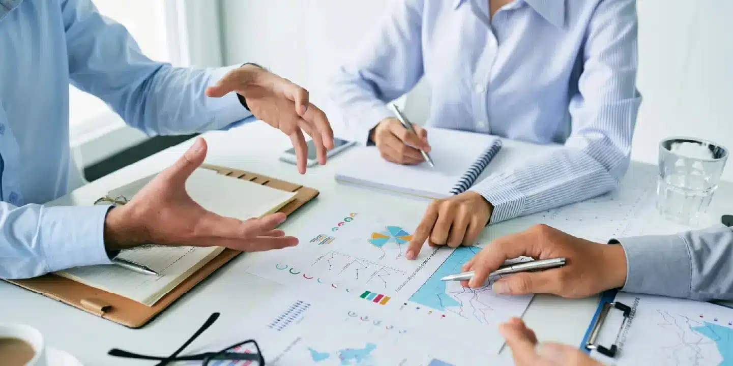 Three people sit at a table reviewing documents and charts, taking notes, and discussing data on recent PRO.FILE PLM Updates in a business meeting setting.