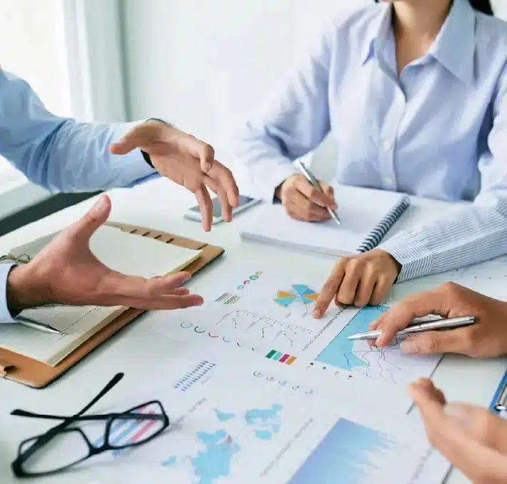 Three people in business attire discuss financial charts and reports at a table, surrounded by notebooks, papers, eyeglasses, and pens while reviewing recent PRO.FILE PLM Updates.