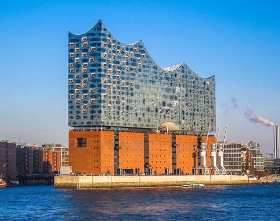 Eine moderne gläserne Konzerthalle mit einem gewellten Dach steht auf einem historischen Backsteinlagerhaus am Fluss in einer Stadtlandschaft unter einem klaren blauen Himmel.