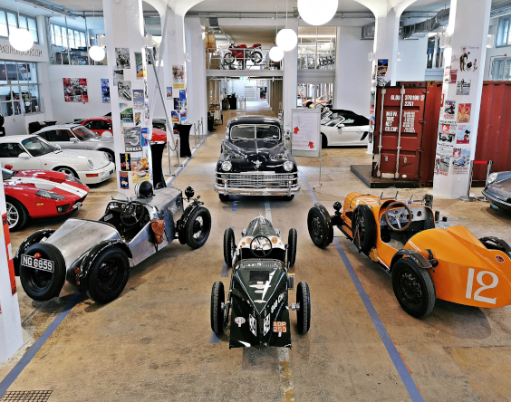 In einem überdachten Ausstellungsraum mit weißen Säulen und Postern an den Wänden ist eine Sammlung von Oldtimern und Klassikern ausgestellt.