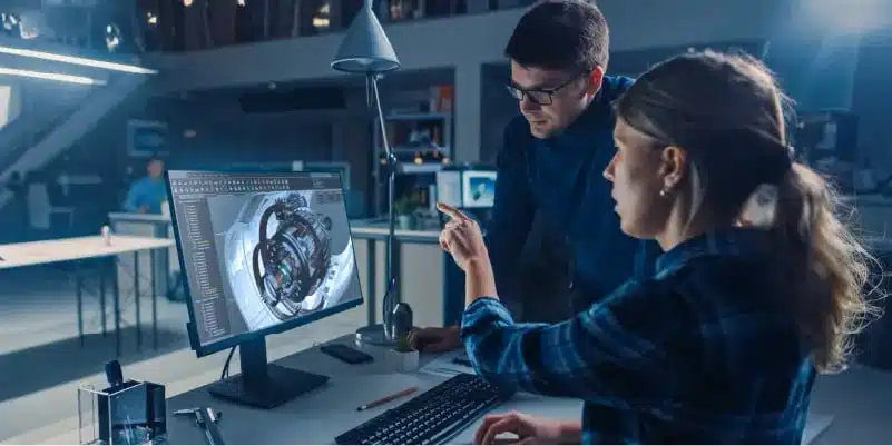 Two people in an office review a 3D engine model displayed on a computer screen, discussing the technical design in a modern workspace.
