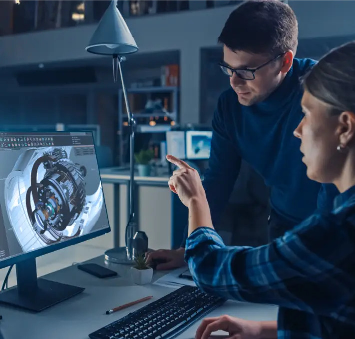 Two people at a desk analyze a 3D model on a computer screen in an office environment, with one person pointing at the monitor.