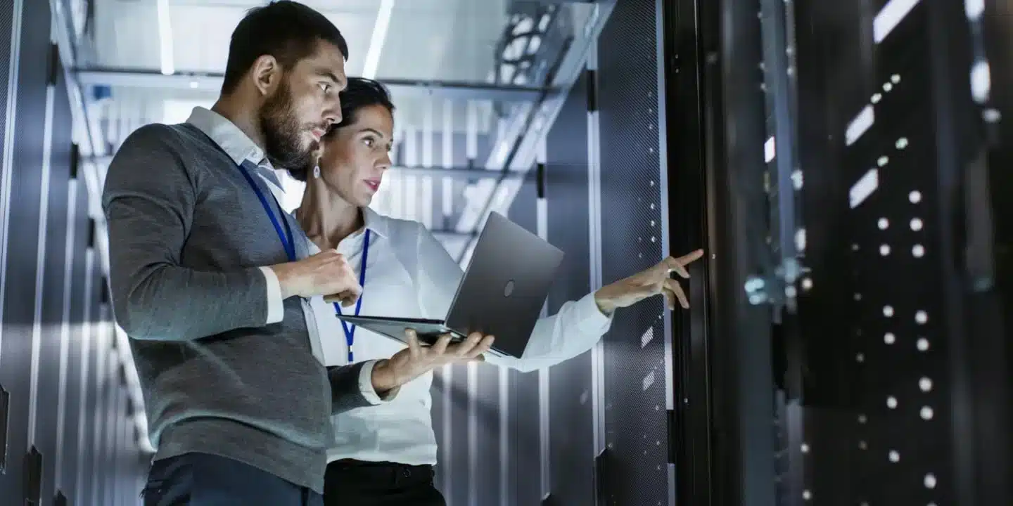 Two IT professionals inspect server racks in a data center; one holds a laptop while the other points at equipment.