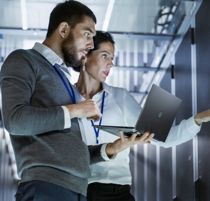 Two professionals examine equipment in a server room, one holding a laptop and the other pointing at a server rack.
