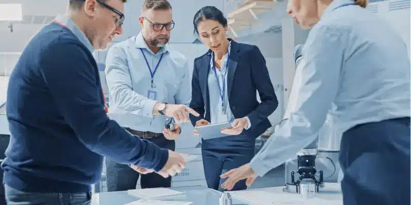 Vier Fachleute in Geschäftskleidung stehen in einem Büro um einen Tisch herum, besprechen Dokumente und benutzen ein Tablet.