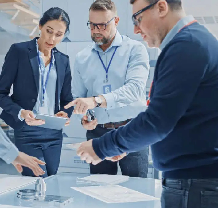 Three people in business attire review documents and digital information together at a desk in a modern office setting.