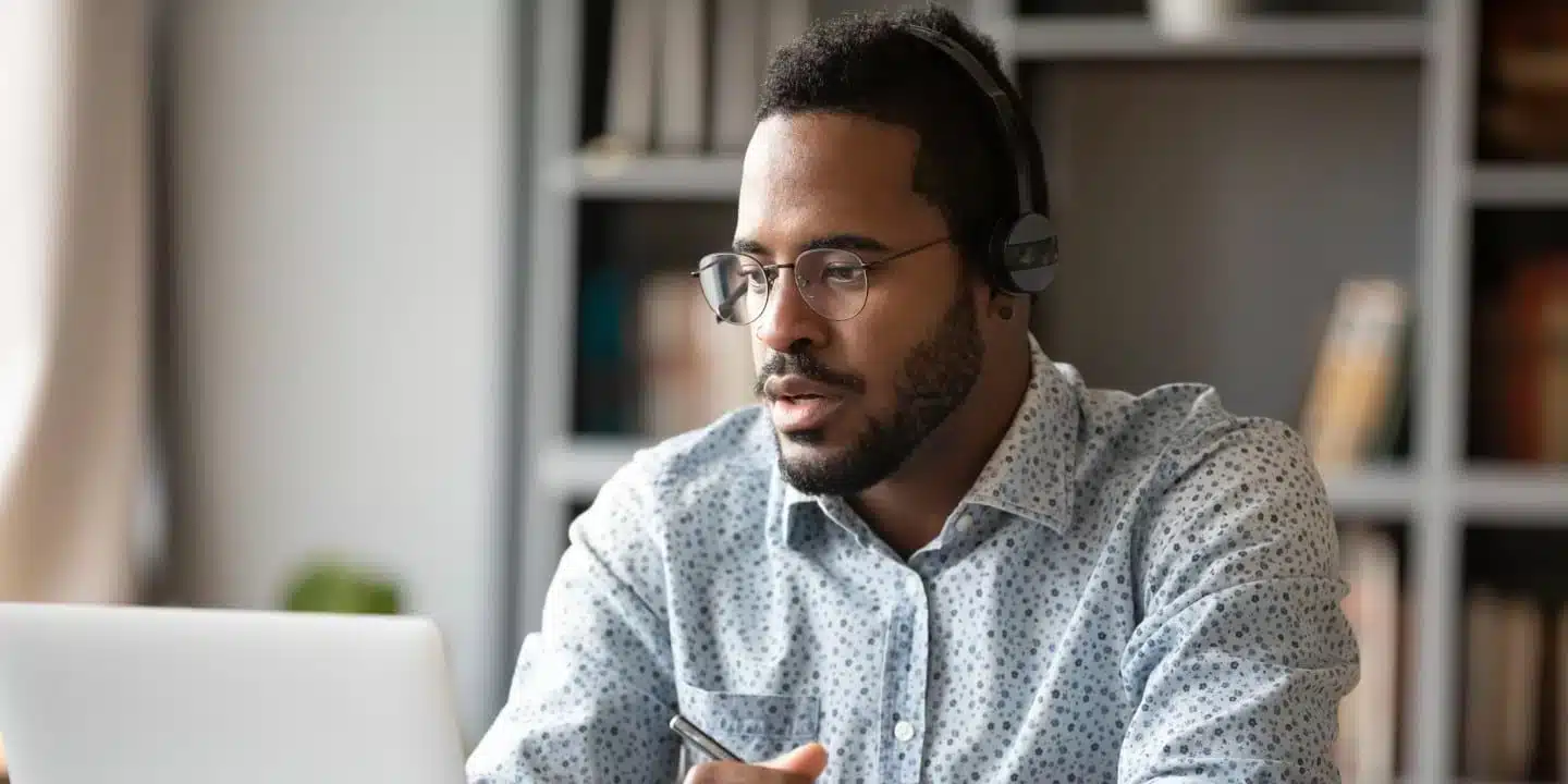 Ein Mann mit Kopfhörern und Brille sitzt vor einem Laptop und scheint konzentriert zu arbeiten, in einem Raum mit Bücherregalen im Hintergrund.