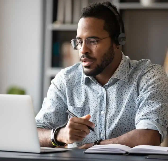 A man wearing glasses and headphones sits at a desk, looking at a laptop screen, holding a pen, with an open notebook in front of him.