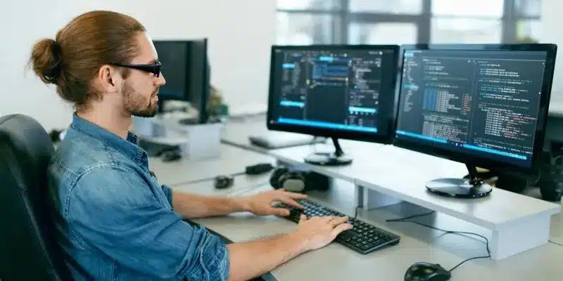 A person with glasses and a ponytail works at a desk with two monitors displaying code in an office setting.