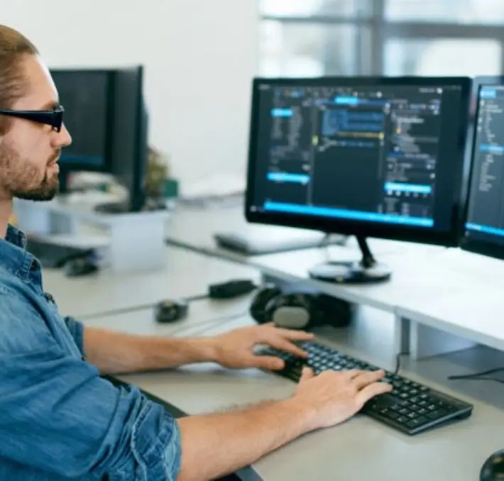 A man with glasses and a ponytail is sitting at a desk, typing on a keyboard and looking at a computer monitor displaying code in an office setting.