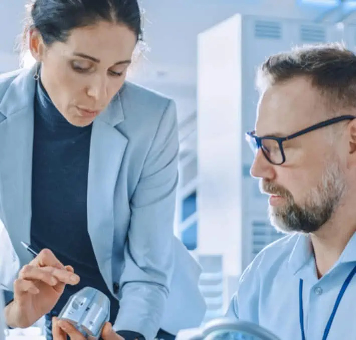 Two professionals in business attire examine a small electronic device in an office or laboratory setting, discussing how dms software solutions can streamline their workflow.
