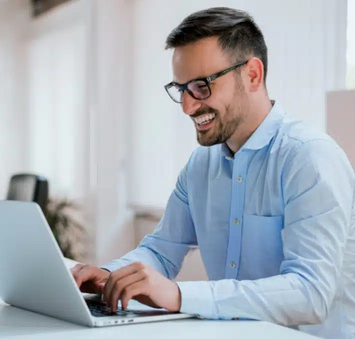 A man wearing glasses and a blue shirt is smiling while working on a laptop with dms software solutions at a desk in a bright office setting.