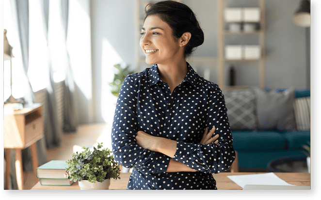 Woman in a polka dot shirt stands indoors with arms crossed, smiling and looking out a window; she feels confident with strong profile support, while a plant and books are on the desk in front of her.