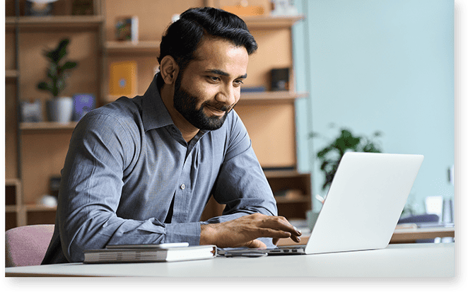 Man in a gray shirt working on a laptop at a desk with a notebook, providing profile support in a modern office setting.