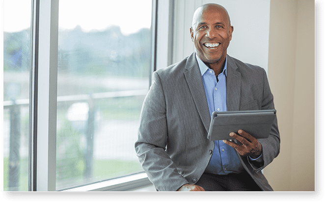 A man in a suit sits by a window, smiling and holding a tablet, ready to offer profile support.