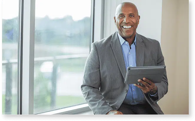 A man in a suit sits by a window, smiling and holding a tablet, ready to offer profile support.