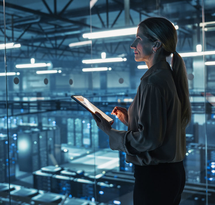 A woman stands in a modern data center, holding a tablet and looking at server racks behind a glass wall.