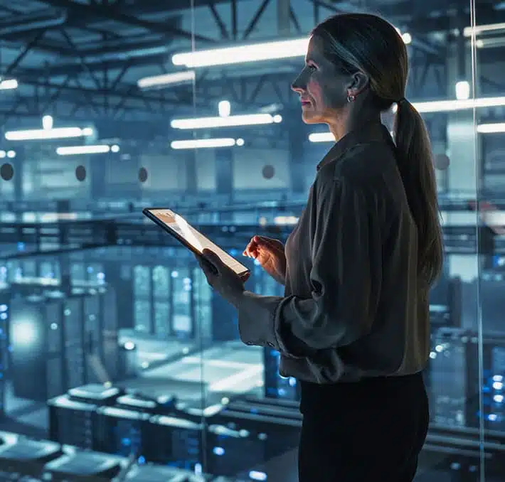 A woman holding a tablet stands in a modern industrial facility, overseeing rows of equipment and illuminated workspaces, highlighting the importance of understanding PDM vs PLM in managing complex manufacturing processes.