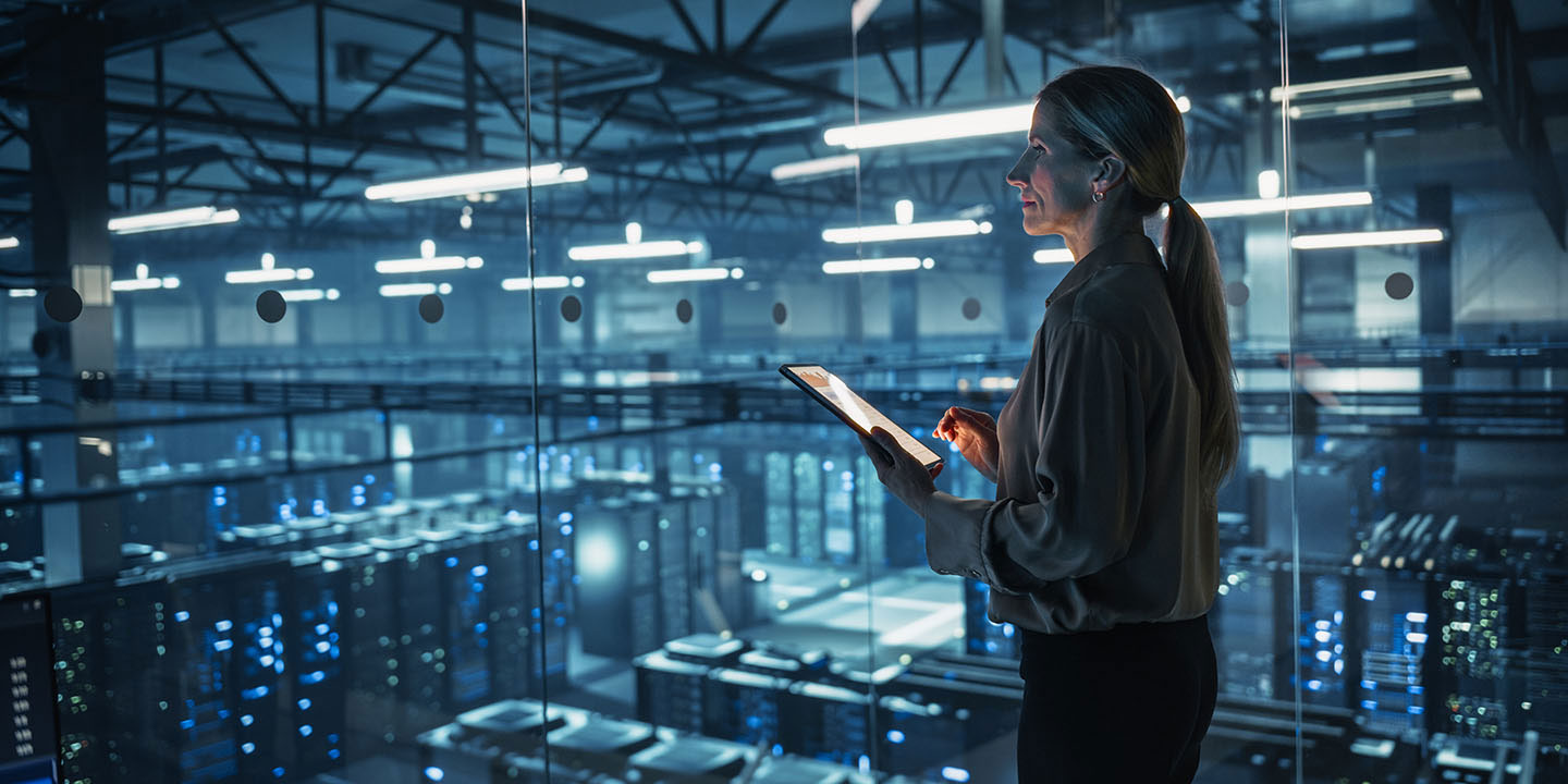 A woman holding a tablet stands in a glass-walled room overlooking a large, modern data center filled with server racks.