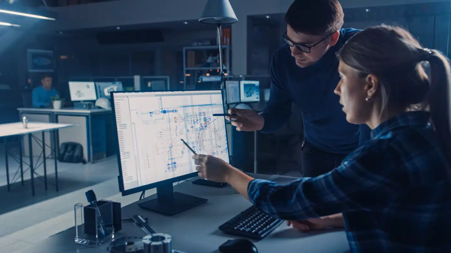 Two people review technical blueprints on a computer monitor in a modern office setting, discussing and pointing at details on the screen.