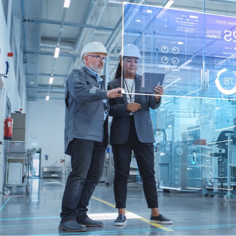 Two engineers wearing safety helmets stand in a modern factory, reviewing data on a tablet as digital interface graphics—highlighting pdm vs plm features—are projected in front of them.