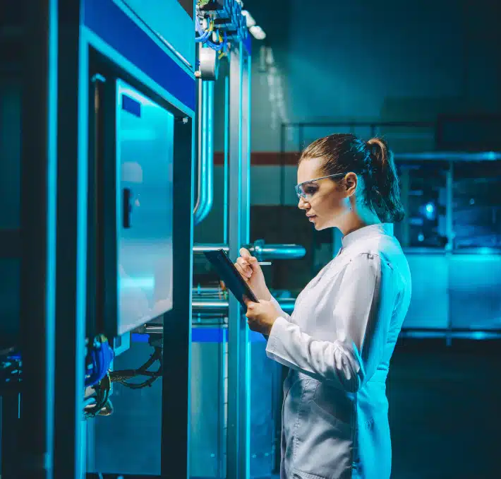 A woman in a lab coat and safety glasses writes on a clipboard, utilizing web-based document management while standing next to industrial equipment in a laboratory or factory setting.