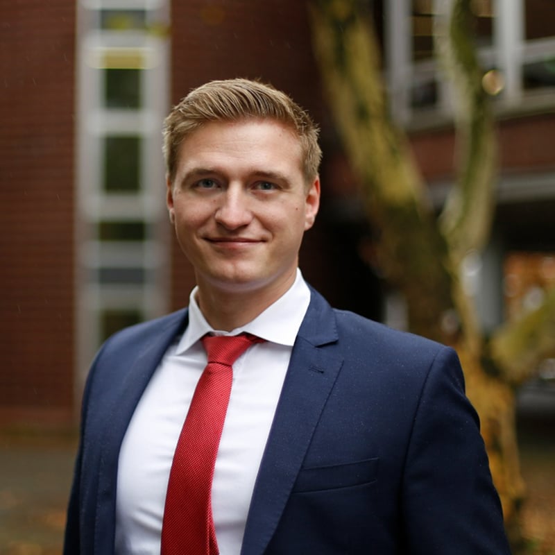 A man in a blue suit and red tie stands outdoors, smiling confidently, with a blurred building and tree in the background—embodying the professionalism and reliability associated with a strong PLM data foundation.