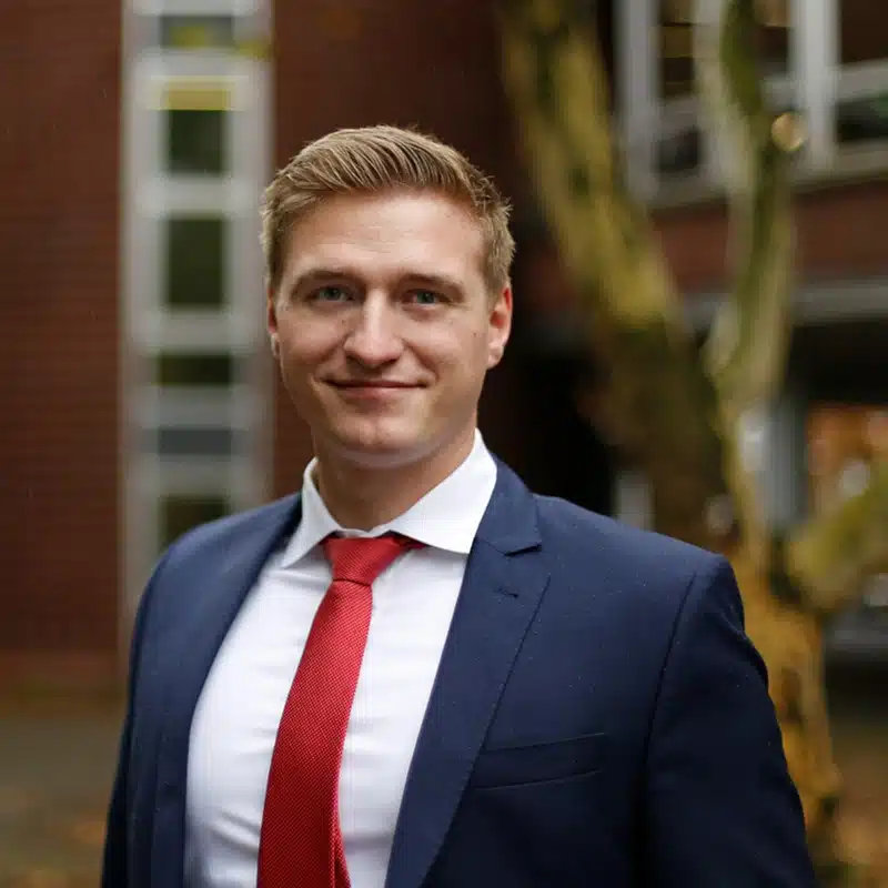 A man in a blue suit and red tie stands outdoors, smiling confidently, with a blurred building and tree in the background—embodying the professionalism and reliability associated with a strong PLM data foundation.