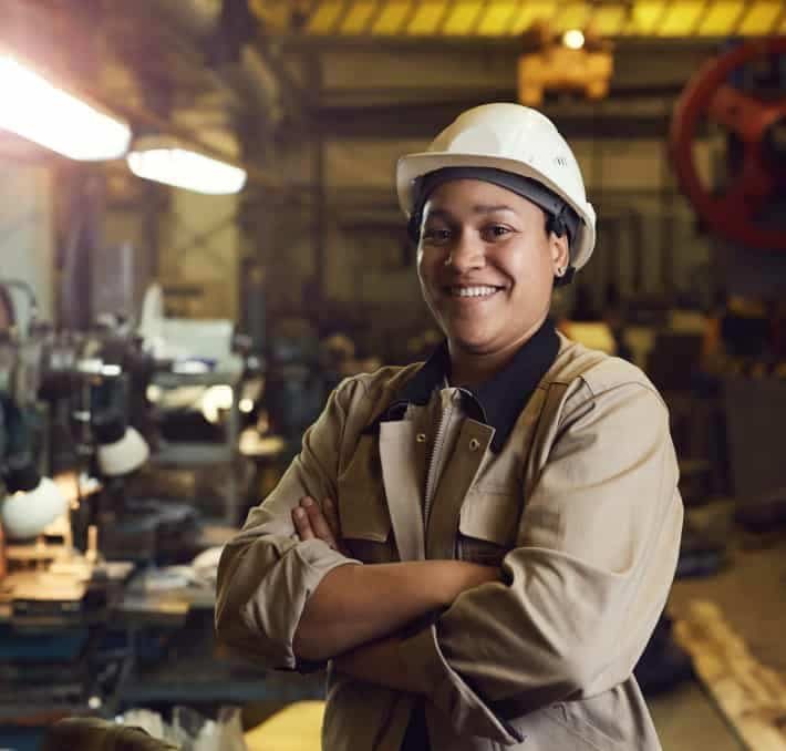 A person wearing a white hard hat and beige work jacket stands with arms crossed, smiling in an industrial workshop setting focused on efficient BOM management.