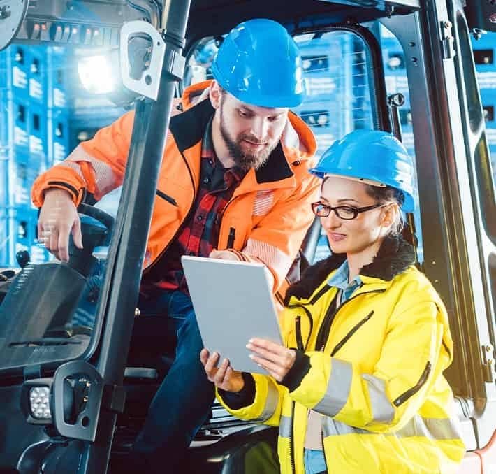 Two warehouse workers in safety gear and blue helmets review information on a tablet next to industrial shelving.