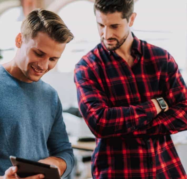 Two men standing indoors; one in a blue shirt holding a tablet and smiling, while his PLM partner in a red plaid shirt looks at the tablet with a serious expression.