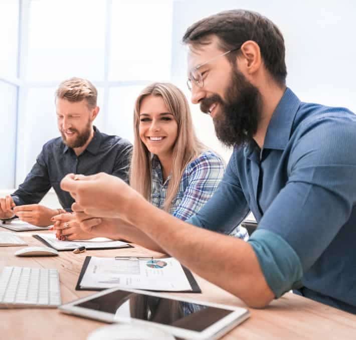 Three people sit at a desk with documents, a keyboard, and a tablet, discussing dms software solutions during a work meeting in a bright office setting.