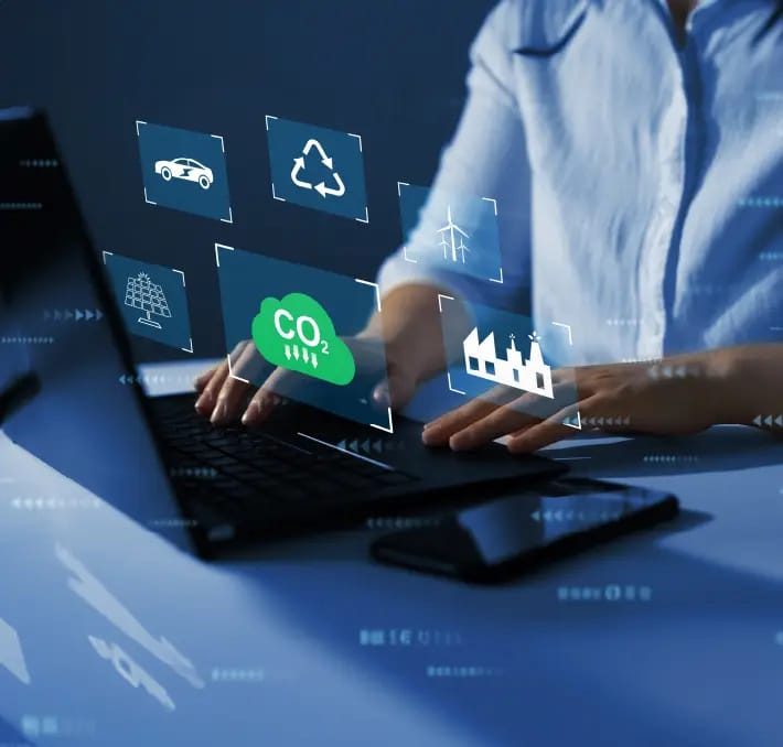 Person typing on a laptop with digital icons overlayed, including symbols for CO2, recycling, car, solar energy, wind energy, industry, and Product Data Management.