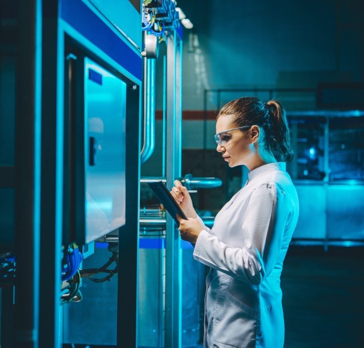 A woman in a lab coat and safety glasses writes on a clipboard, utilizing web-based document management while standing next to industrial equipment in a laboratory or factory setting.